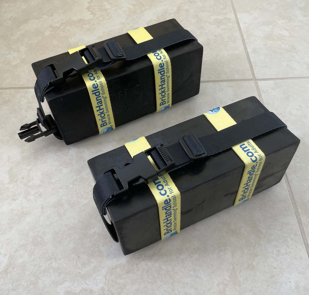 two black diving bricks, each with a yellow and black buckled handle to carry it